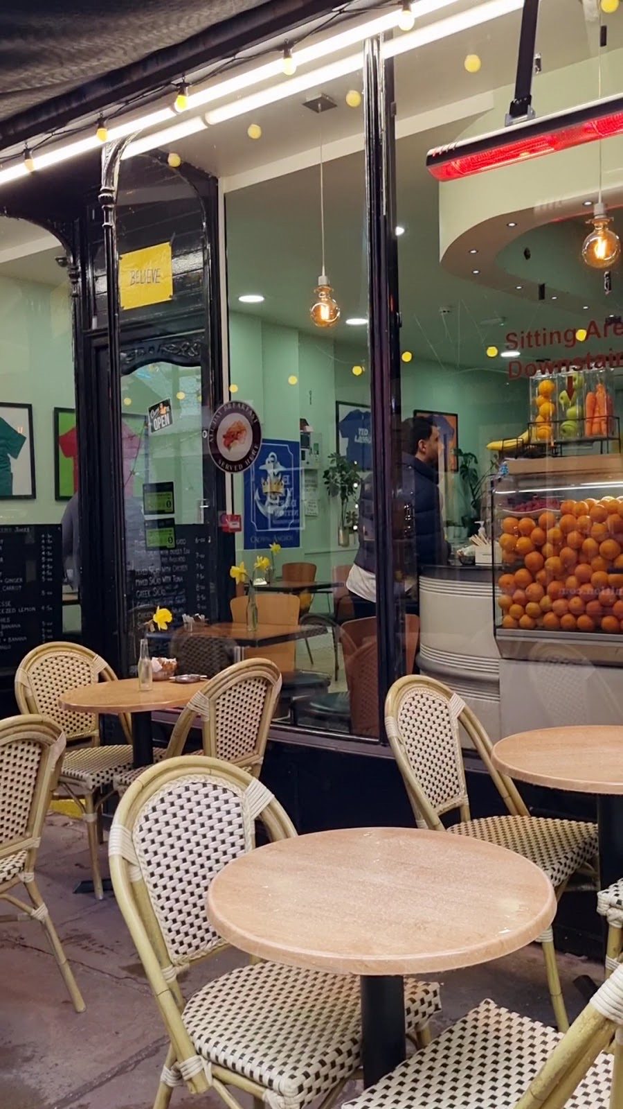 Inside the café looking out through the shopfront window, pendant lights over the tables and a fruit display by the counter
