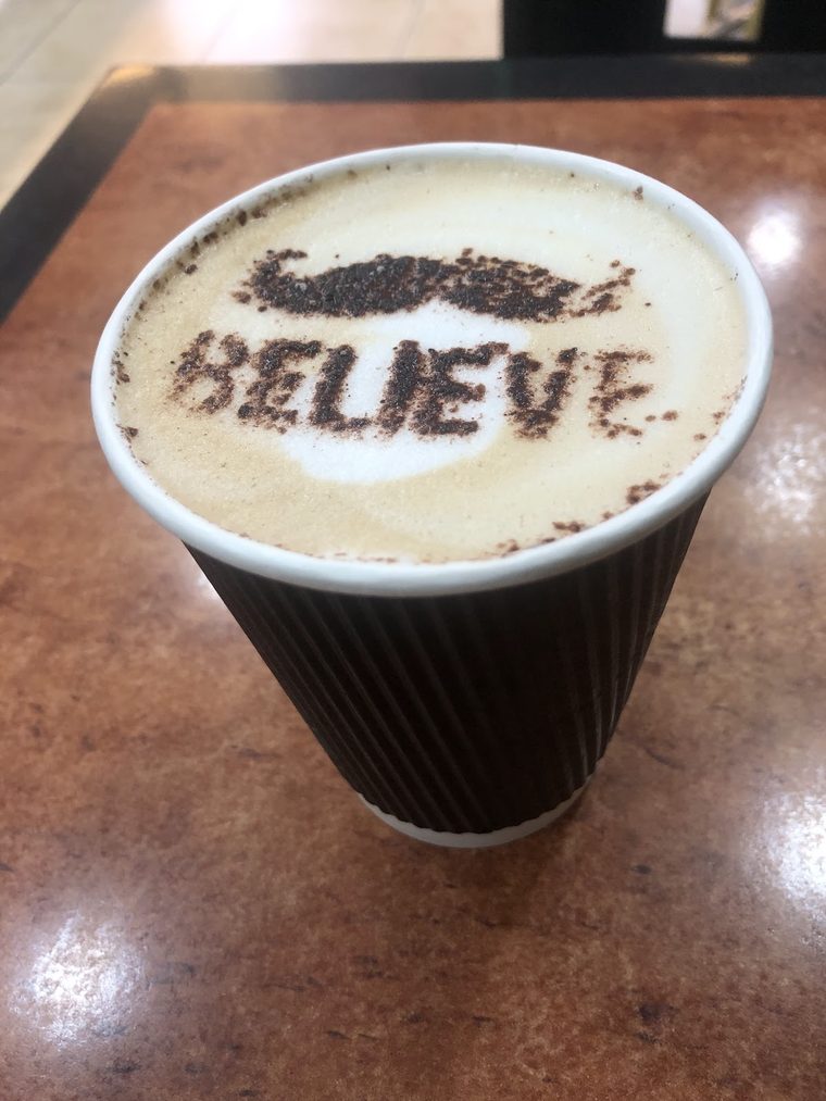 A takeaway coffee cup with cocoa stencilled on the foam, on a wooden table, cropped tight on the cup to leave the lettering out